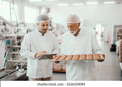 Senior adult employee holding tray with fresh cookies while supervisor evaluating quality and holding tablet. Both are dressed in sterile white uniforms and having hairnets. Food plant interior. - Powered by Shutterstock