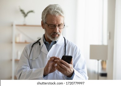 Senior Adult Male Doctor Holding Smart Phone Using Remote Medical Consultation Applications, Texting Messages, Chatting With Distance Patient Online Using Telemedicine Mobile App Standing In Hospital.