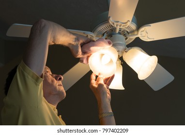 Senior Adult Male Dusting The Glass Shade Of A Bulb In A Ceiling Fan And Lighting Fixture