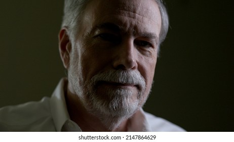 A senior adult man in a dimly lit room smiles a little smile for the camera.