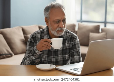 A SENIOR ADULT MAN DRINKING TEA WHILE LOOKING AT LAPTOP