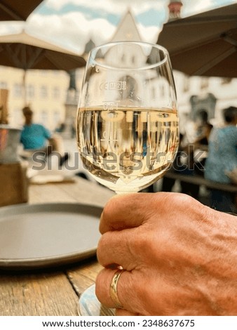 Senior adult man holds the stem of a wine glass full of white wine. The man is sitting at an outdoor cafe or pub in a quaint European town square. CE translates to European Conformity.