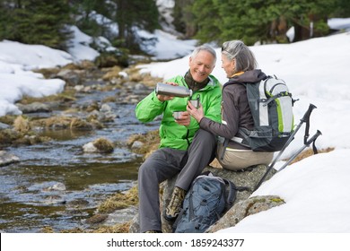 A senior adult man pouring coffee for his wife while taking a small break from trekking at the edge of a stream in the snowy woods