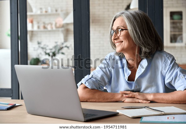 Senior adult mid 50s age watching at window at
home sitting at table with laptop. Feeling happy and smiling to
thoughts about future positive vision of successful training career
after learning.