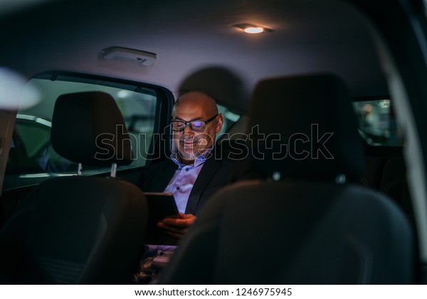 Senior adult sitting on backseat and using tablet\
at night.