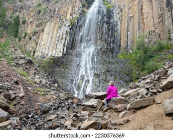 Senior adult woman wearing a fuchsia jacket and gray pants sitting on a boulder at the base of Palisade Falls in Hyalite Canyon, Montana, on an overcast day. - Shutterstock ID 2230376369