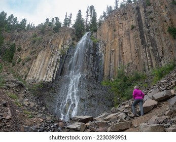 Senior adult woman wearing a fuchsia jacket and gray pants standing at the base of Palisade Falls in Hyalite Canyon, Montana, on an overcast day. - Shutterstock ID 2230393951