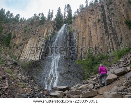 Senior adult woman wearing a fuchsia jacket and gray pants standing at the base of Palisade Falls in Hyalite Canyon, Montana, on an overcast day.