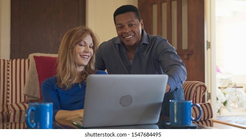 Senior African American And Caucasian Husband And Wife Use Laptop Computer Together In Living Room. Older Homeowner Married Couple Smiling While Booking Trip Online