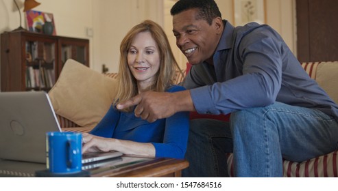 Senior African American And Caucasian Husband And Wife Use Laptop Computer Together In Living Room. Older Homeowner Married Couple Smiling While Booking Trip Online