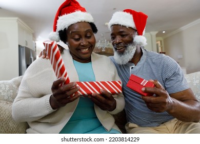 Senior African American Couple Opening A Gift, Smiling And Wearing Santa Hats. Quality Family Time Christmas Celebration.