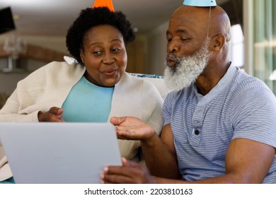 Senior African American Couple Spending Time At Home Together, Sitting On Sofa, Wearing Party Hats, Using A Laptop Computer. Isolating During Coronavirus Covid 19 Quarantine Lockdown.