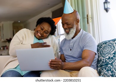 Senior African American Couple Spending Time At Home Together, Sitting On Sofa, Wearing Party Hats, Using A Laptop Computer. Isolating During Coronavirus Covid 19 Quarantine Lockdown.