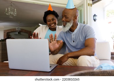 Senior African American Couple Spending Time At Home Together, Sitting On Sofa, Wearing Party Hats, Using A Laptop Computer. Isolating During Coronavirus Covid 19 Quarantine Lockdown.