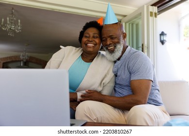 Senior African American Couple Spending Time At Home Together, Sitting On Sofa, Wearing Party Hats, Using A Laptop Computer. Isolating During Coronavirus Covid 19 Quarantine Lockdown.