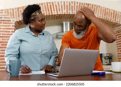 Senior African American Couple Spending Time At Home Together, Standing In Kitchen, Using A Laptop Computer And Looking At Paperwork. Isolating During Coronavirus Covid 19 Quarantine Lockdown.