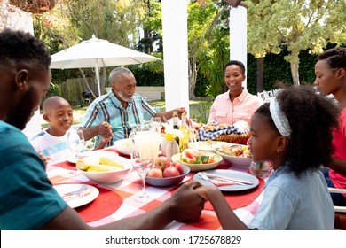 Senior African American Couple And Their Family Spending Time Together In The Garden On A Sunny Day, Sitting By A Table And Praying Before Eating Lunch.