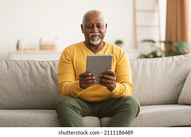 Senior African American Male Using Digital Tablet Reading Online Book And Browsing Internet Sitting On Sofa At Home. Mature Man Holding Computer Relaxing In Living Room. Older People And Gadgets