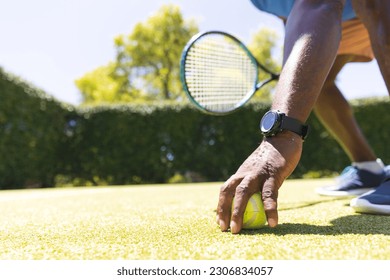 Senior african american man bending down to pick up tennis ball on sunny grass court. Senior lifestyle, retirement, sport, summer, fitness, hobbies and leisure activities. - Powered by Shutterstock