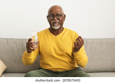 Senior African American Man Holding Medical Pill And Glass Of Water Looking At Camera Sitting On Sofa Indoors. Medicine Treatment, Vitamins And Supplements In Older Age Concept