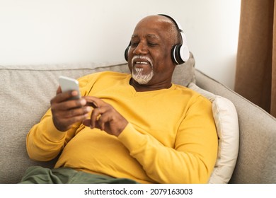 Senior African American Man Listening To Music On Phone Wearing Headphones Sitting On Sofa At Home, Relaxing On Weekend. Older People Using Gadgets, Technology And Modern Lifestyle