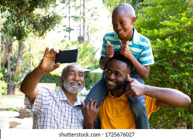 Senior African American Man Spending Time With His Son And His Grandson In The Garden On A Sunny Day, Taking A Selfie With A Smartphone.