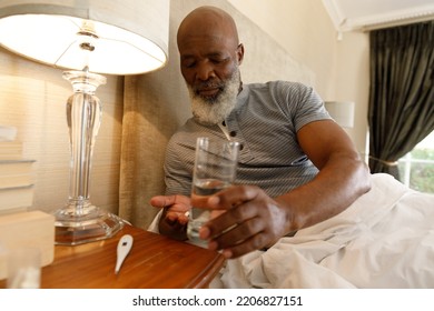 Senior African American Man Spending Time At Home, Lying In Bed, Holding Glass Of Water And Taking Medication. Isolating During Coronavirus Covid 19 Quarantine Lockdown.