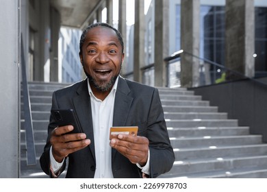 A senior African American man in a suit stands outside an office. He is holding a credit card and a phone. Successfully paid online purchases, shopping, orders, bills. Happy looking at the camera.