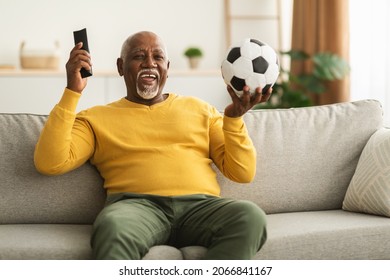 Senior African American Man Watching Sport On TV Celebrating Victory Of Favorite Team Holding Ball And Remote Controller Sitting At Home. Grandfather Watches Soccer Game On Television On Weekend