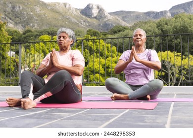 Senior African American woman and senior biracial woman are practicing yoga outdoors. They are seated on yoga mats, performing a pose with a serene mountain backdrop. - Powered by Shutterstock