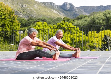 Senior African American woman and senior biracial woman are smiling while doing yoga outdoors. They are stretching on yoga mats, surrounded by lush greenery, promoting health and wellness. - Powered by Shutterstock