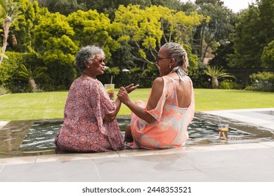 Senior African American woman and senior biracial woman enjoy a conversation by the pool. They are dressed in summer attire, sharing a moment in a lush garden setting. - Powered by Shutterstock