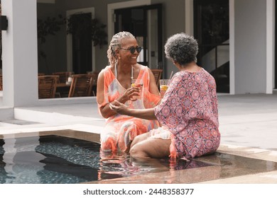 Senior African American woman and senior biracial woman enjoy a conversation by the poolside. Both are dressed in vibrant summer attire, reflecting a leisurely day in a serene outdoor setting. - Powered by Shutterstock