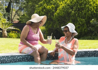 Senior African American woman and senior biracial woman enjoy drinks by the pool. They are wearing sun hats and smiling, basking in a sunny, outdoor setting. - Powered by Shutterstock