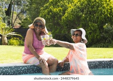 Senior African American woman and senior biracial woman toast drinks by a pool. Both wear sun hats and sunglasses, enjoying a sunny day outdoors. - Powered by Shutterstock