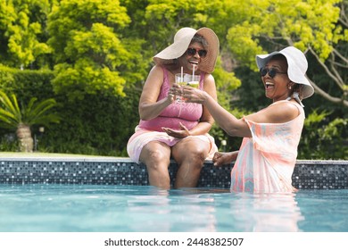 Senior African American woman and senior biracial woman enjoy drinks in a pool with copy space. They are laughing together, wearing sun hats and summer dresses, embodying leisure and friendship. - Powered by Shutterstock