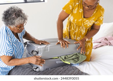 Senior African American woman and senior biracial woman pack a suitcase together. They are preparing for a trip, organizing clothes in a bedroom setting. - Powered by Shutterstock