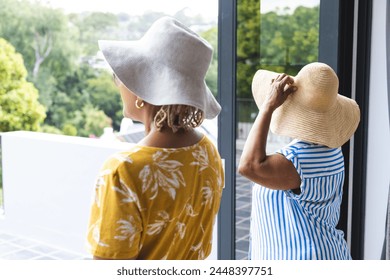 Senior African American woman and senior biracial woman enjoy a view from a balcony on vacation. Both wear wide-brimmed hats and summer dresses, embracing a leisurely day. - Powered by Shutterstock