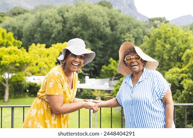 Senior African American woman and senior biracial woman share a joyful moment outdoors on vacation. Both wear sun hats and sunglasses, exuding happiness in a lush, green setting. - Powered by Shutterstock