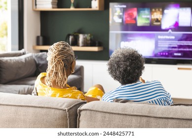 Senior African American woman and senior biracial woman are sitting on a couch, watching TV at home. They are relaxed in a cozy living room, enjoying leisure time together. - Powered by Shutterstock