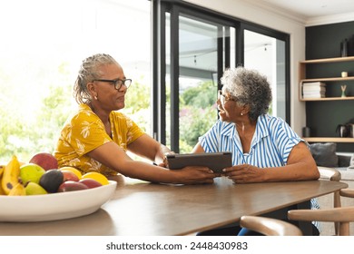 Senior African American woman and senior biracial woman sit at a table, sharing a tablet at home. They are engaged in a conversation, surrounded by a bright, modern kitchen setting. - Powered by Shutterstock