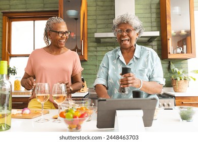 Senior African American woman and senior biracial woman share a joyful moment in a kitchen. They are laughing together, surrounded by cooking ingredients and a tablet, suggesting a cooking session. - Powered by Shutterstock