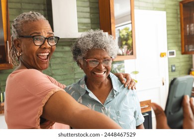 Senior African American woman and senior biracial woman share a joyful moment taking a selfie. They are in a kitchen setting, radiating happiness with wide smiles and casual attire. - Powered by Shutterstock