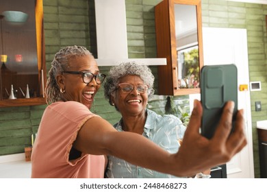 Senior African American woman and senior biracial woman are taking a selfie with a phone. Both are smiling joyfully, capturing a moment of friendship in a cozy kitchen setting. - Powered by Shutterstock