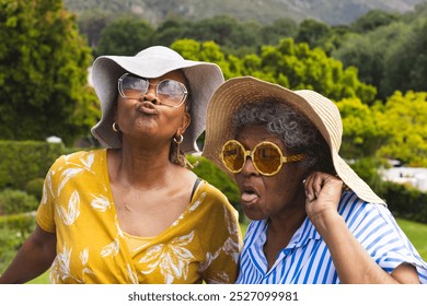 Senior African American woman and biracial woman make playful faces, wearing sun hats, on vacation. Their joyful expressions and outdoor setting suggest a lighthearted moment during a leisurely day. - Powered by Shutterstock