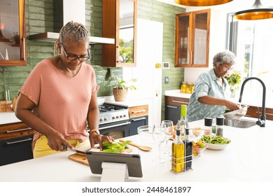 Senior African American woman chops vegetables as a senior biracial woman washes produce. They are preparing a meal together in a bright, modern kitchen, showcasing teamwork and healthy living. - Powered by Shutterstock