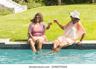 Senior African American woman enjoys a laugh with a friend by the poolside, both holding drinks. They are dressed in vibrant summer attire, reflecting a leisurely day outdoors. - Powered by Shutterstock