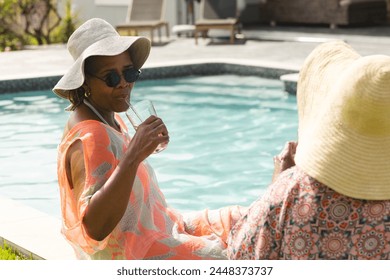 Senior African American woman sips a drink by the pool, wearing a sunhat and sunglasses. Senior biracial woman faces her, both enjoying a leisurely conversation outdoors. - Powered by Shutterstock