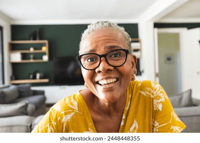 Senior African American woman smiles warmly on video call, wearing glasses and a yellow top. Her joyous expression brightens the cozy home setting. - Powered by Shutterstock