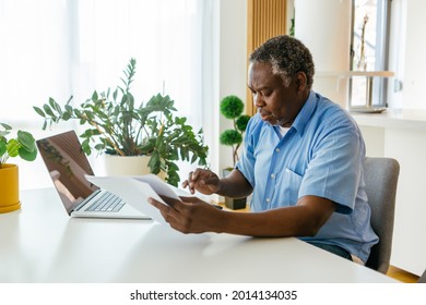 Senior African man sitting in his home office with bills in hands and paying it online over laptop. E banking concept.
